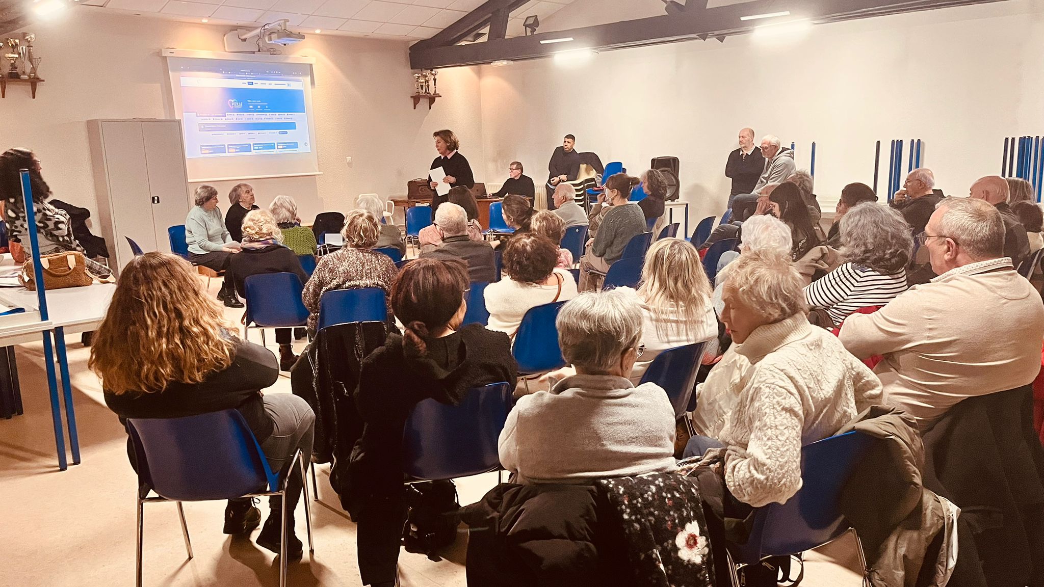 Réunion publique du 31 janvier — Catherine au pupitre, site VeM projeté, salle pleine