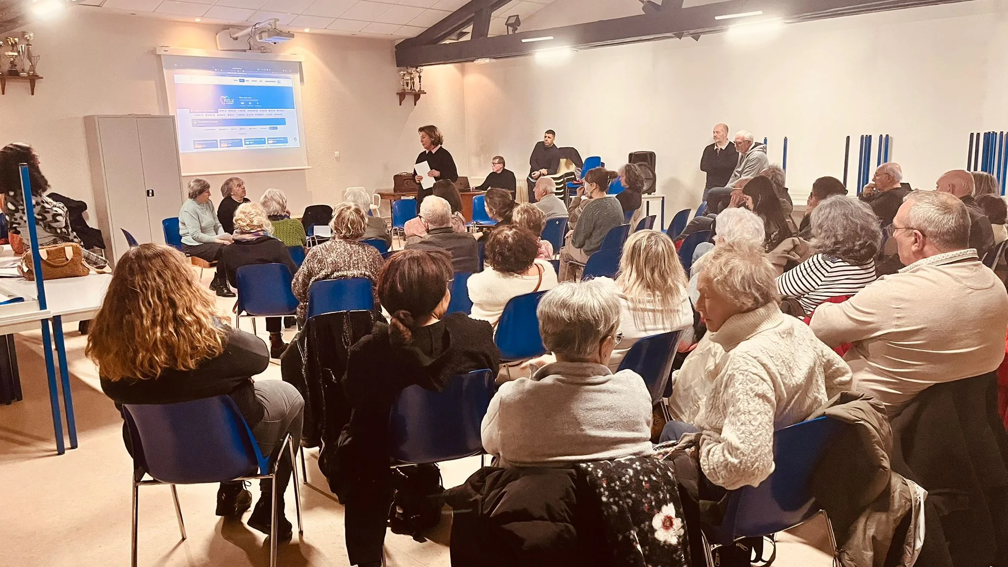 Réunion publique du 31 janvier — Catherine au pupitre, site VeM projeté, salle pleine
