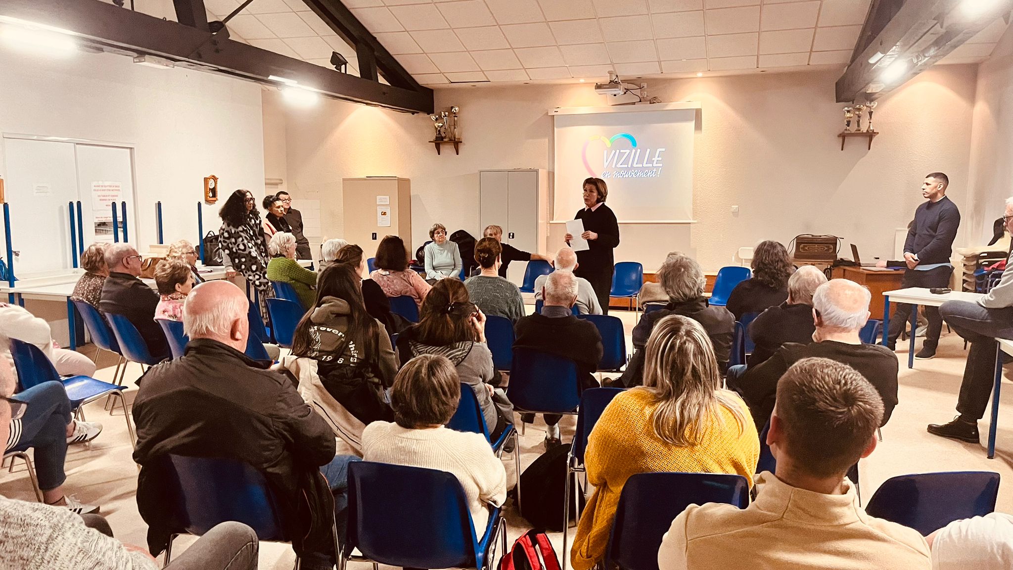 Vue d'ensemble — salle comble, logo Vizille en Mouvement projeté