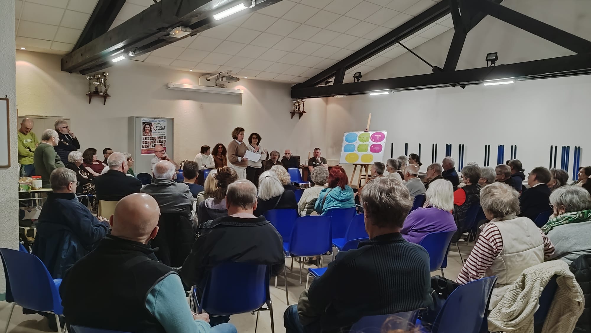 Grande réunion du 27 février — salle pleine, tableau programme coloré, affiche Catherine