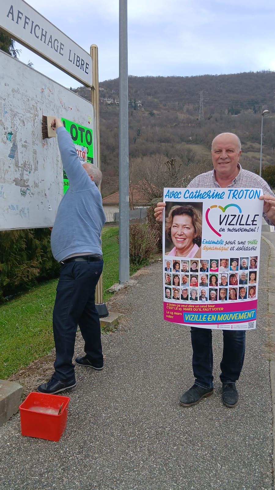 Pose d'affiches — un candidat tient l'affiche VeM, l'autre nettoie le panneau